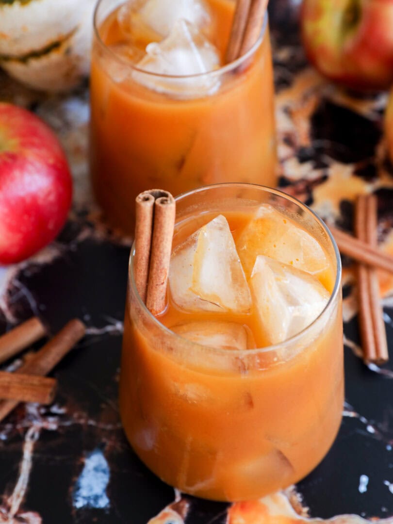 Two glasses of iced apple cider garnished with cinnamon sticks are on a marble surface, surrounded by fresh apples, loose cinnamon sticks for a cozy autumn twist.