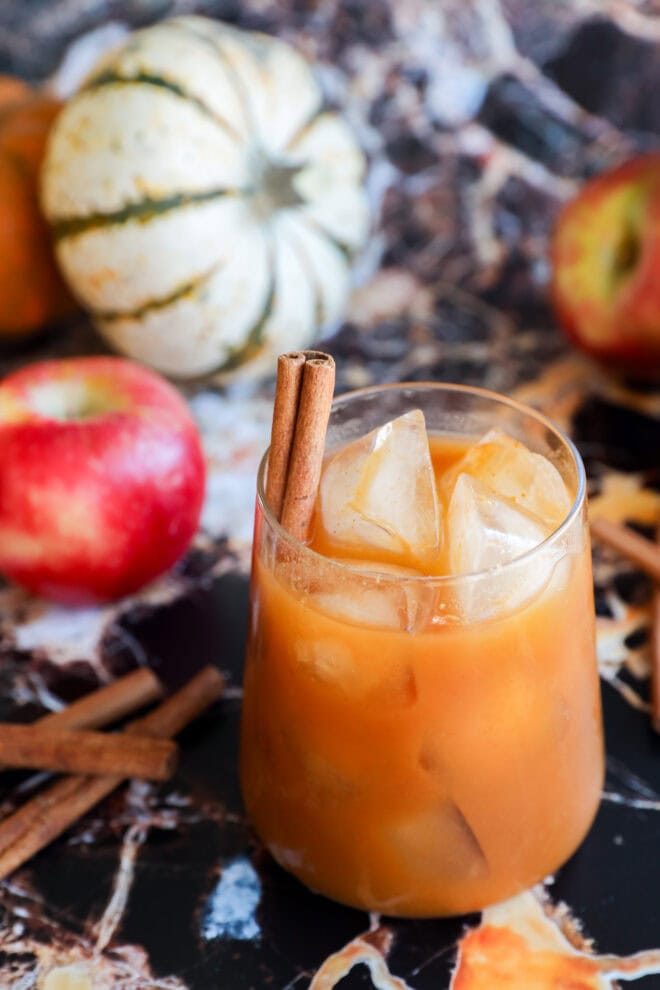 A glass of orange iced pumpkin juice garnished with two cinnamon sticks, surrounded by apples, extra cinnamon sticks, and small decorative pumpkins on a marble surface.