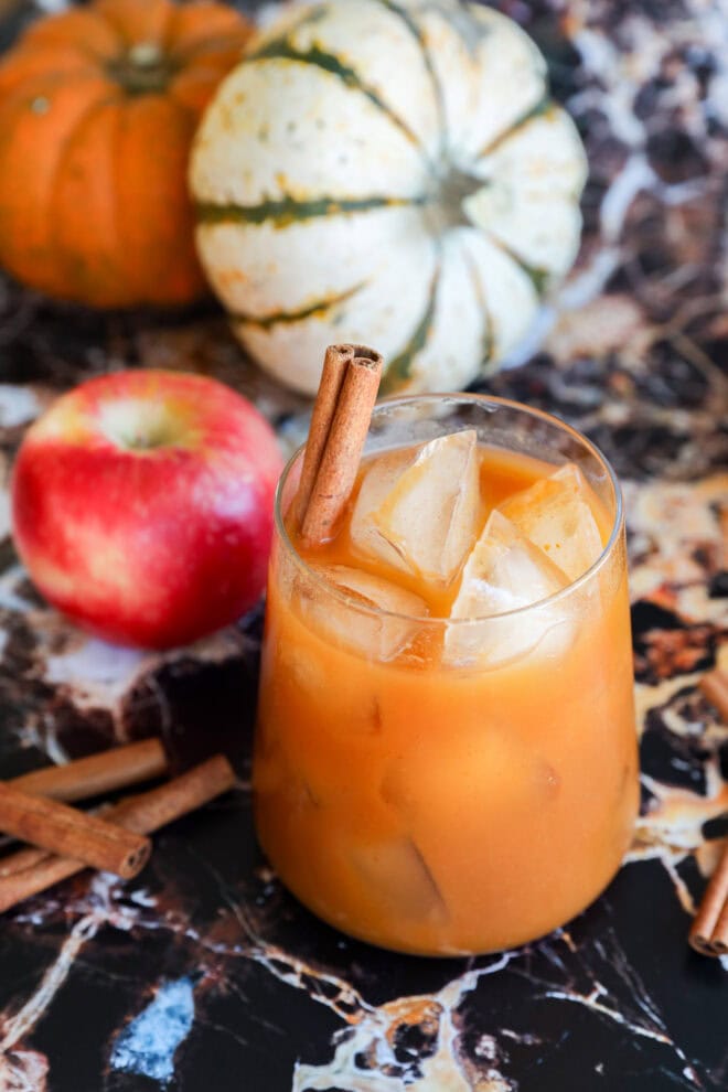 A glass of orange-colored iced pumpkin juice with a cinnamon stick, set on a dark marbled surface, with an apple, cinnamon sticks, and two decorative pumpkins in the background.