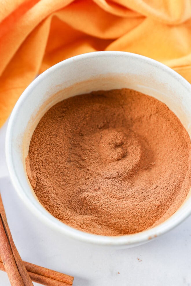 A white bowl filled with a mound of ground cinnamon or pumpkin pie spice, with an orange cloth and cinnamon sticks placed nearby on a light surface.