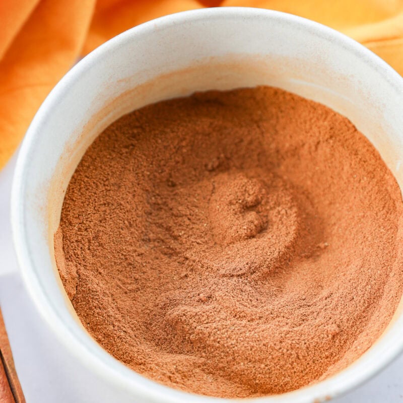 A white bowl filled with a mound of ground cinnamon or pumpkin pie spice, with an orange cloth and cinnamon sticks placed nearby on a light surface.