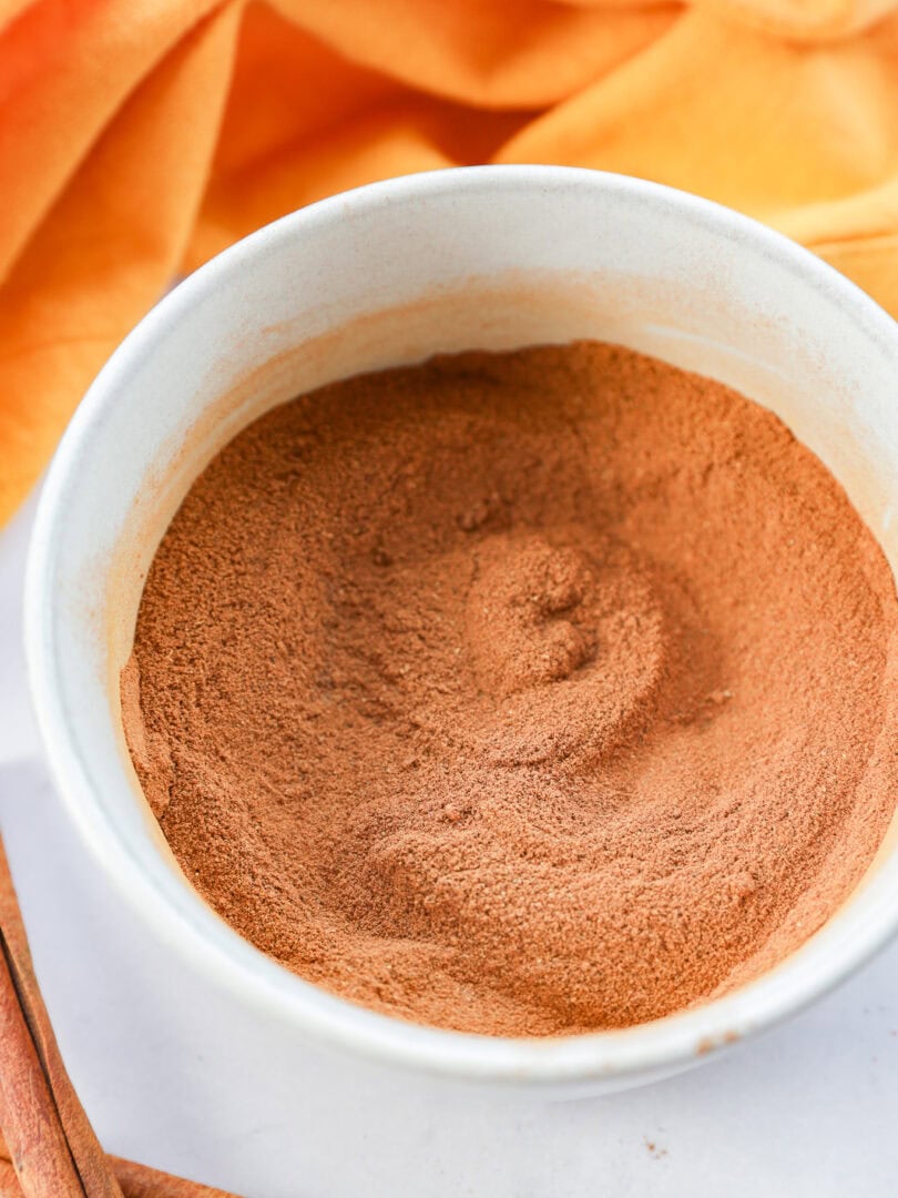 A white bowl filled with a mound of ground cinnamon or pumpkin pie spice, with an orange cloth and cinnamon sticks placed nearby on a light surface.