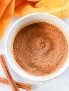 A white bowl filled with ground cinnamon or pumpkin pie spice sits on a white surface. Two cinnamon sticks lie nearby, and an orange cloth is partially visible in the background.