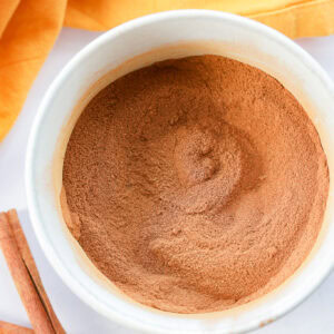 A white bowl filled with ground cinnamon or pumpkin pie spice sits on a white surface. Two cinnamon sticks lie nearby, and an orange cloth is partially visible in the background.
