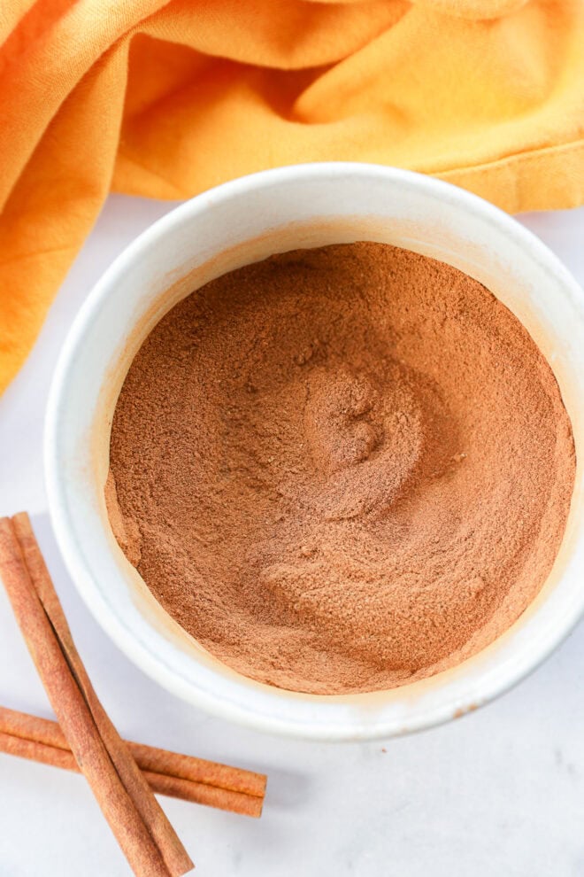 A white bowl filled with ground cinnamon or pumpkin pie spice sits on a white surface. Two cinnamon sticks lie nearby, and an orange cloth is partially visible in the background.