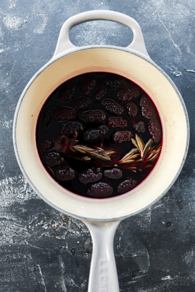A white pot filled with a dark red liquid, blackberries, and star anise, sitting on a textured gray surface.