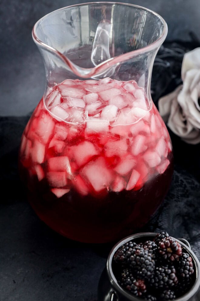 A glass pitcher filled with a red beverage and ice cubes sits on a dark surface. Nearby, a small bowl holds fresh blackberries. A white cloth is partially visible in the background.