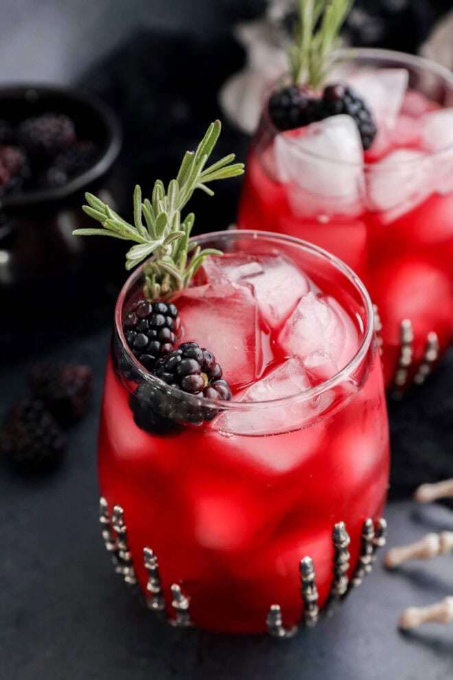 A glass of witches brew over ice, garnished with blackberries and a sprig of rosemary. Another similar drink is in the background, both served in beaded glasses on a dark surface.