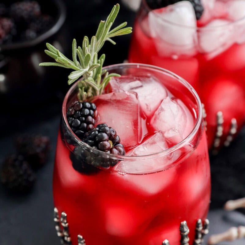 A glass of witches brew over ice, garnished with blackberries and a sprig of rosemary. Another similar drink is in the background, both served in beaded glasses on a dark surface.
