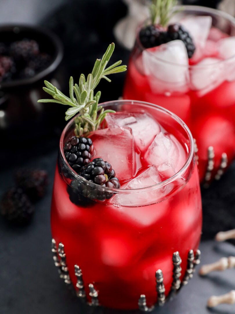 A glass of witches brew over ice, garnished with blackberries and a sprig of rosemary. Another similar drink is in the background, both served in beaded glasses on a dark surface.