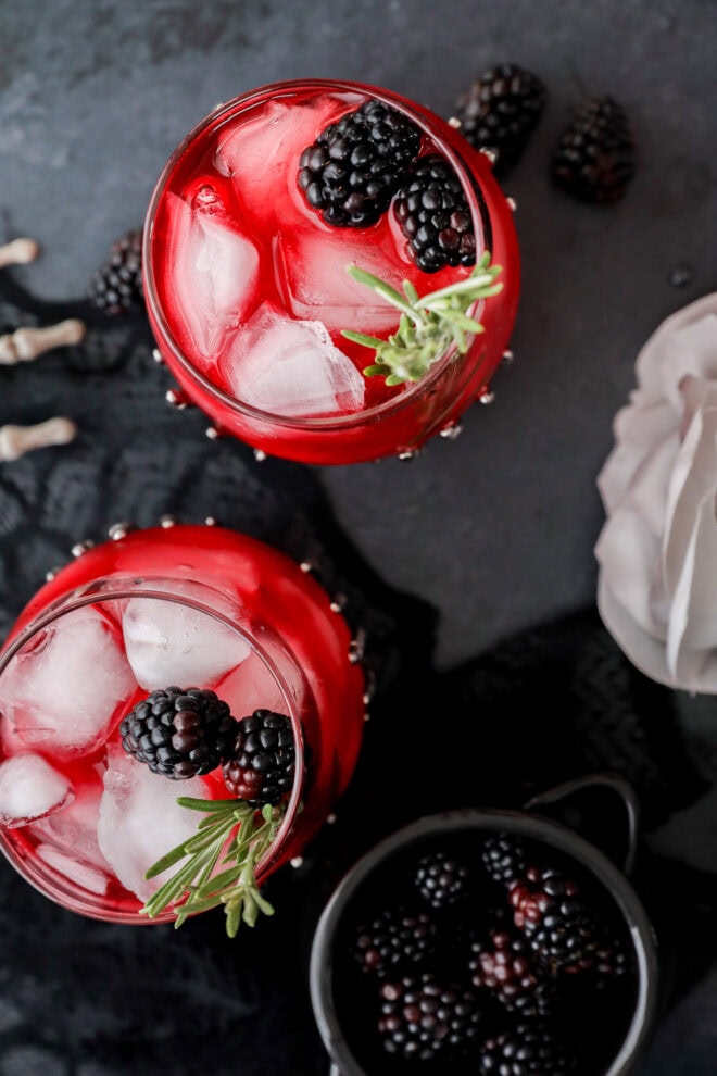A top-down view of a witches brew—a red drink with ice, fresh blackberries, and a sprig of rosemary in a glass, with some blackberries scattered nearby on a dark surface.