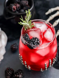 A Witches Brew glass with ice cubes is garnished with blackberries and a rosemary sprig in a decorated glass. A small bowl of blackberries and a skeleton hand prop are in the background, creating a spooky atmosphere.