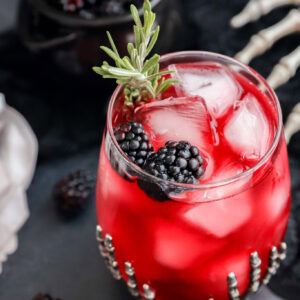 A Witches Brew glass with ice cubes is garnished with blackberries and a rosemary sprig in a decorated glass. A small bowl of blackberries and a skeleton hand prop are in the background, creating a spooky atmosphere.