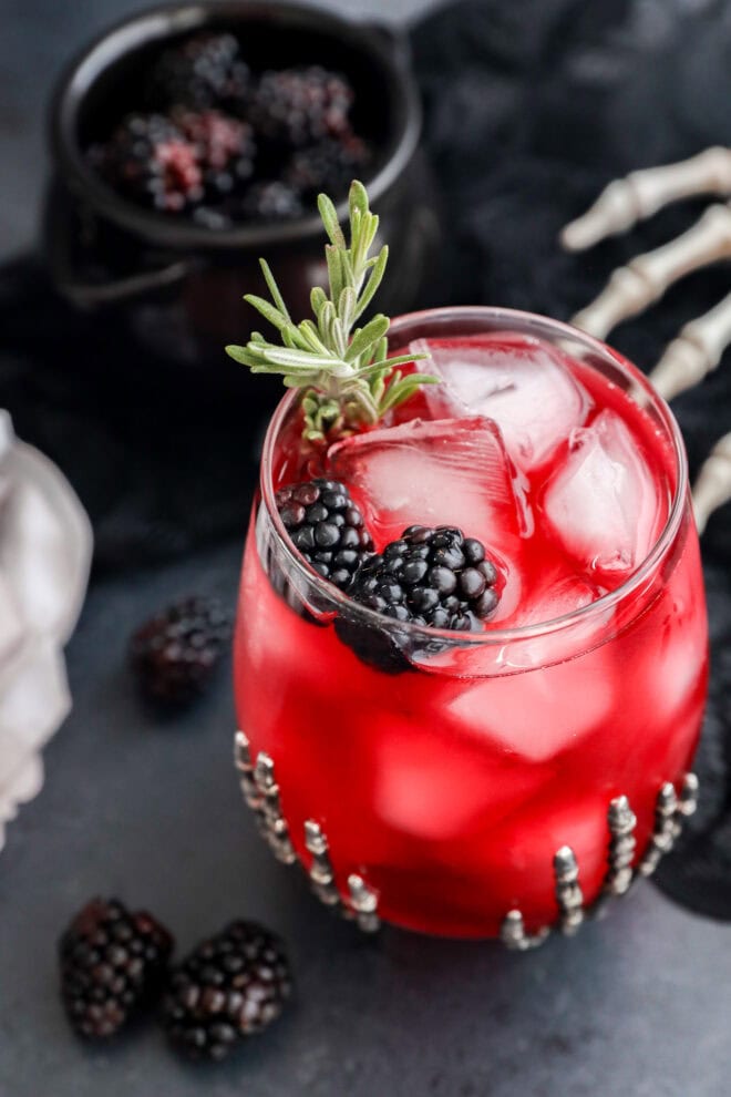 A Witches Brew glass with ice cubes is garnished with blackberries and a rosemary sprig in a decorated glass. A small bowl of blackberries and a skeleton hand prop are in the background, creating a spooky atmosphere.