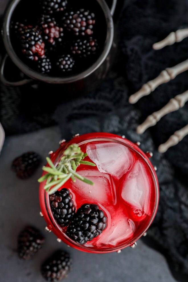A red drink with ice cubes, blackberries, and a sprig of rosemary in a glass, with loose blackberries scattered nearby, a metal bowl of blackberries, and a decorative skeleton hand on a dark surface.