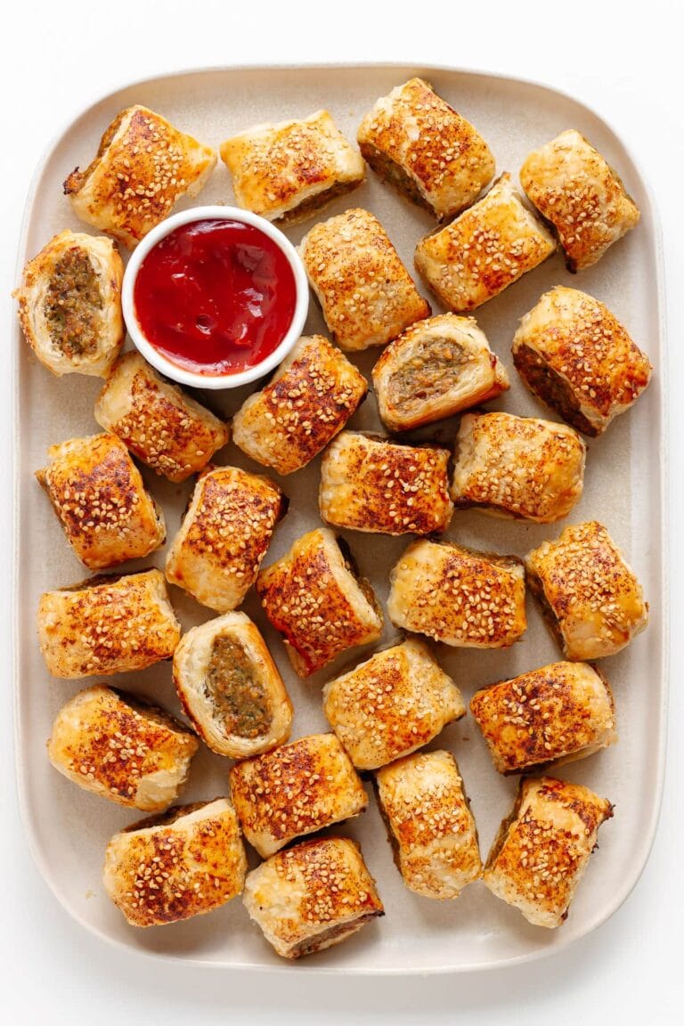 A platter of golden-brown puff pastry appetizers, these sausage rolls are sprinkled with sesame seeds and arranged around a small bowl of red dipping sauce, likely ketchup.