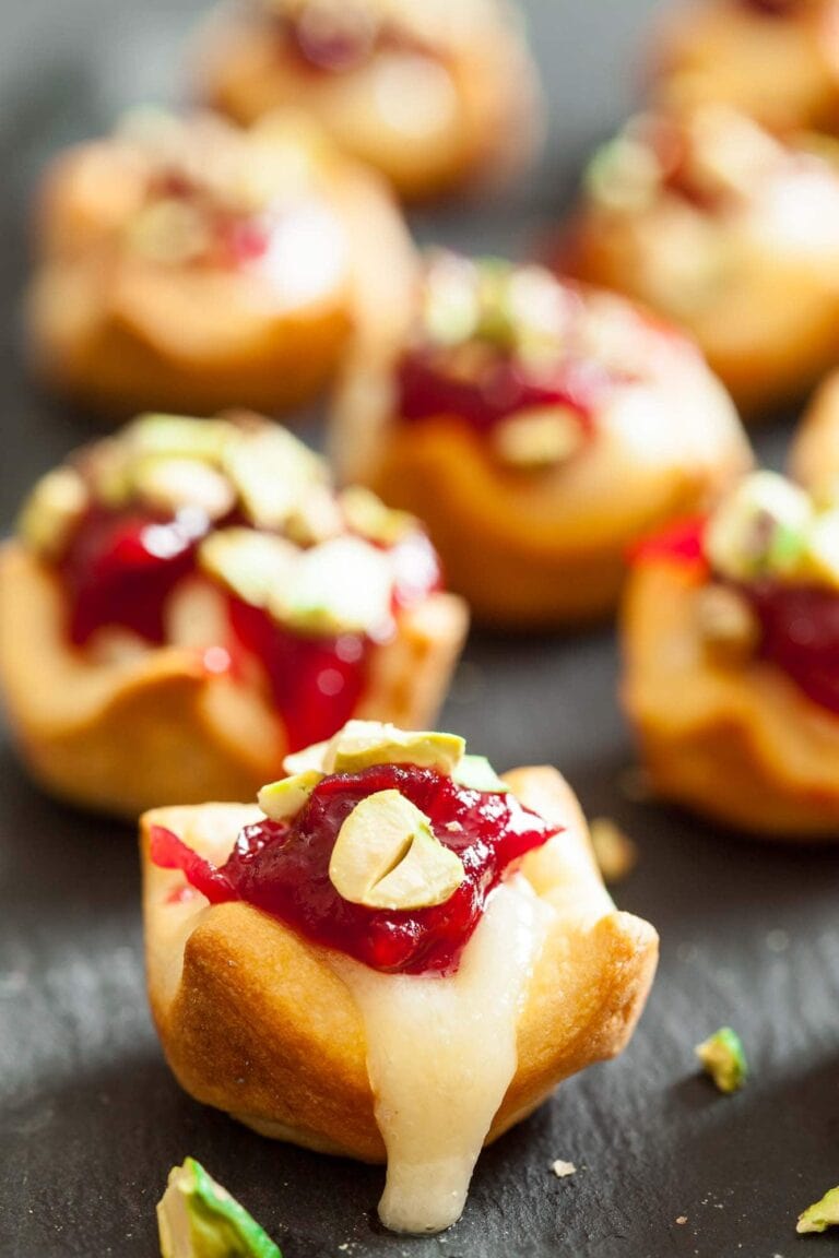 Mini puff pastry appetizers filled with melted cheese, topped with red jelly and chopped green pistachios, arranged on a dark surface.