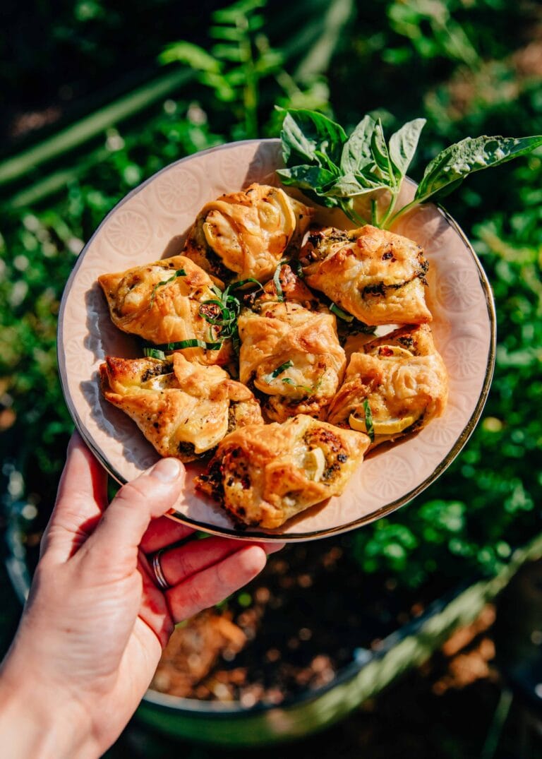 A hand holding a plate with several golden-brown puff pastry appetizers garnished with fresh herbs, with a small bunch of basil on the side, set against a lush green outdoor background.