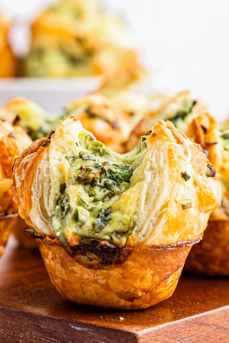 Close-up of golden-brown puff pastry appetizers in a muffin shape, filled with a creamy spinach and herb mixture. The pastry is flaky and layered, with the savory filling visible in the center.