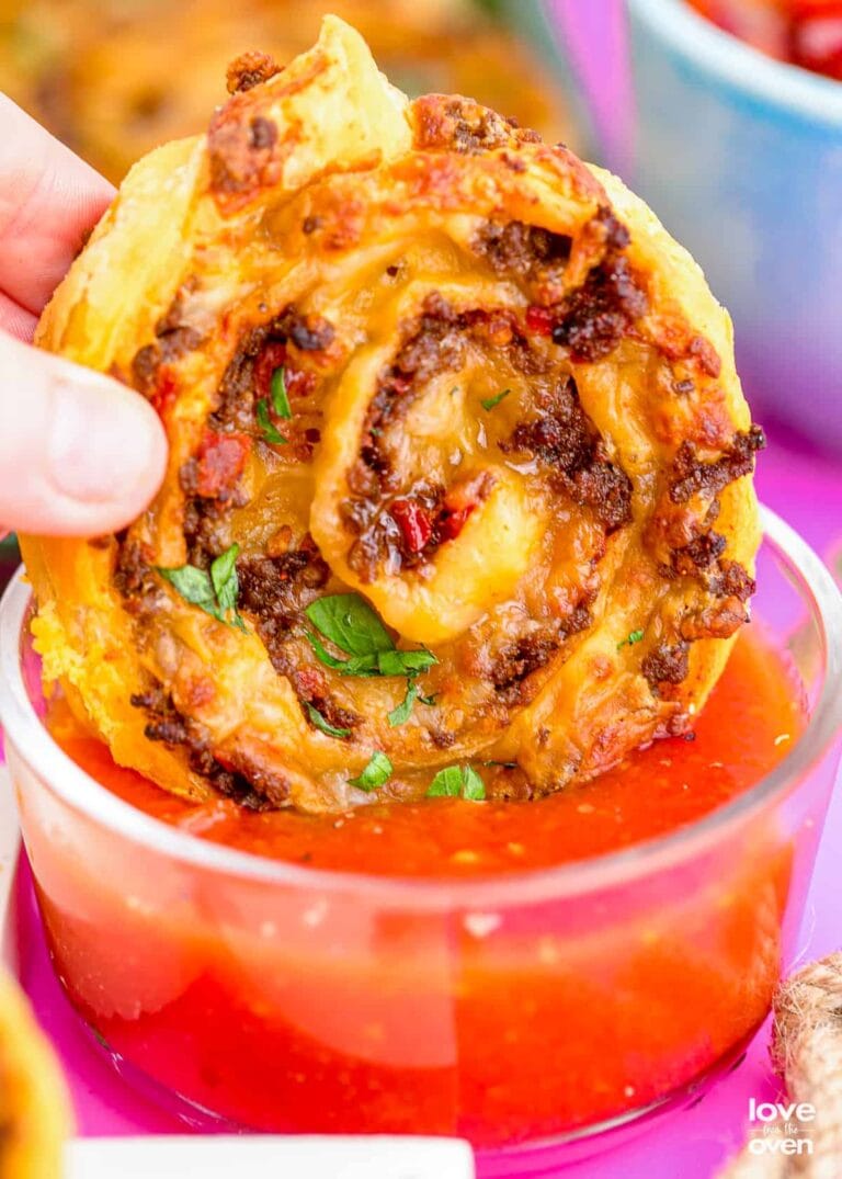 A hand dips a cheesy, spiral-shaped pizza pinwheel—one of the tastiest puff pastry appetizers filled with meat and herbs—into a bowl of marinara sauce.