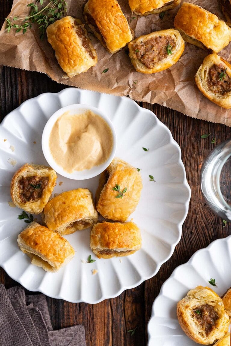 A white plate with several golden-brown puff pastry appetizers, filled as sausage rolls, is served with a creamy dipping sauce and fresh thyme garnish. More savory treats rest on brown parchment paper in the background.