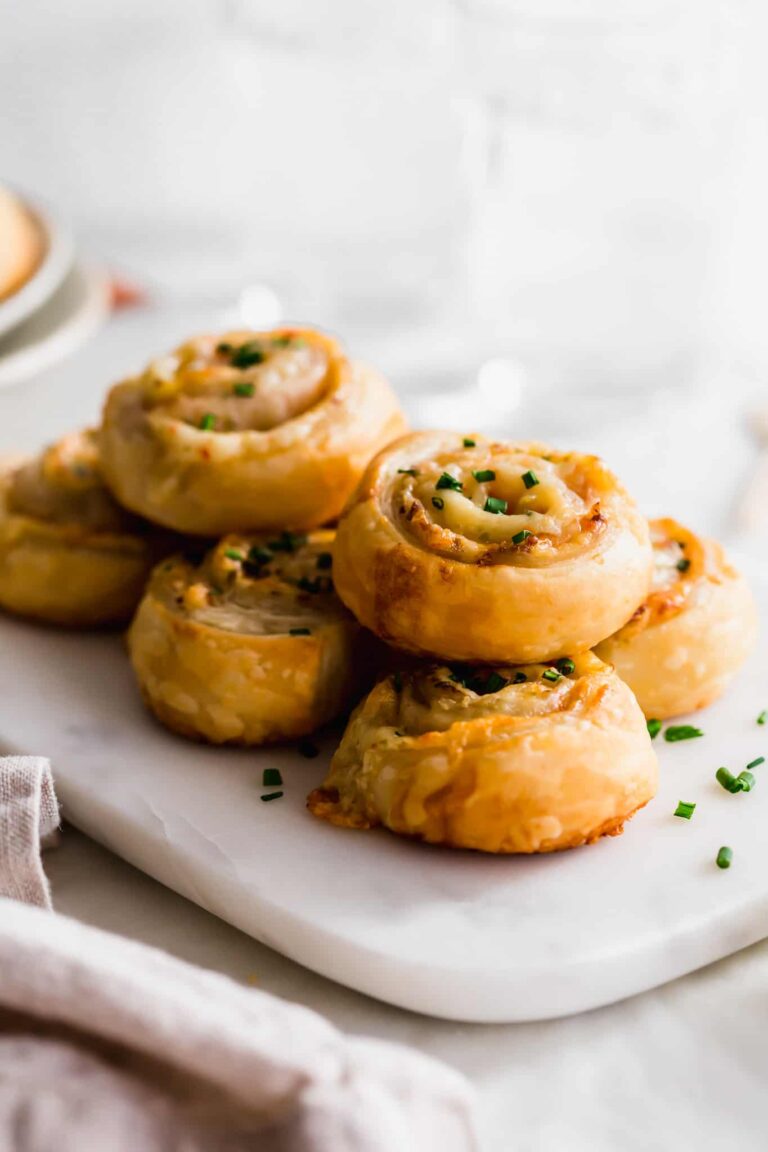 A stack of golden-brown puff pastry appetizers pinwheels, garnished with chopped chives and arranged on a white marble serving board.