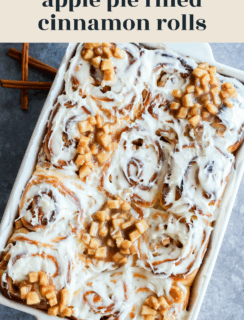 A baking dish filled with Apple Pie Filled Cinnamon Rolls topped with cream cheese icing and chunks of cooked apples, set on a gray surface with cinnamon sticks beside it. Text above reads apple pie filled cinnamon rolls.