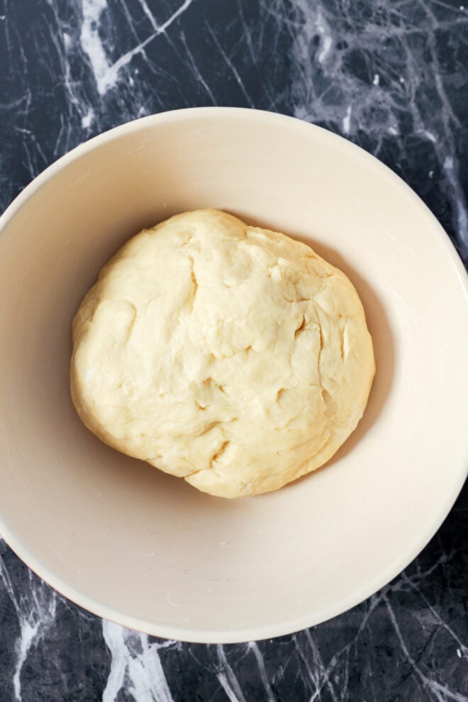 A ball of dough for Apple Pie Filled Cinnamon Rolls rests in a light-colored mixing bowl, placed on a dark, marbled countertop.