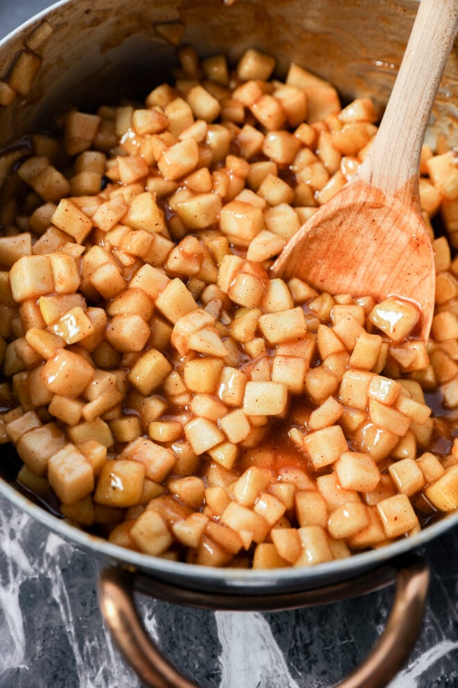A saucepan filled with diced apples coated in a glossy cinnamon sauce, reminiscent of Apple Pie Filled Cinnamon Rolls, is being stirred with a wooden spoon on a marble surface.