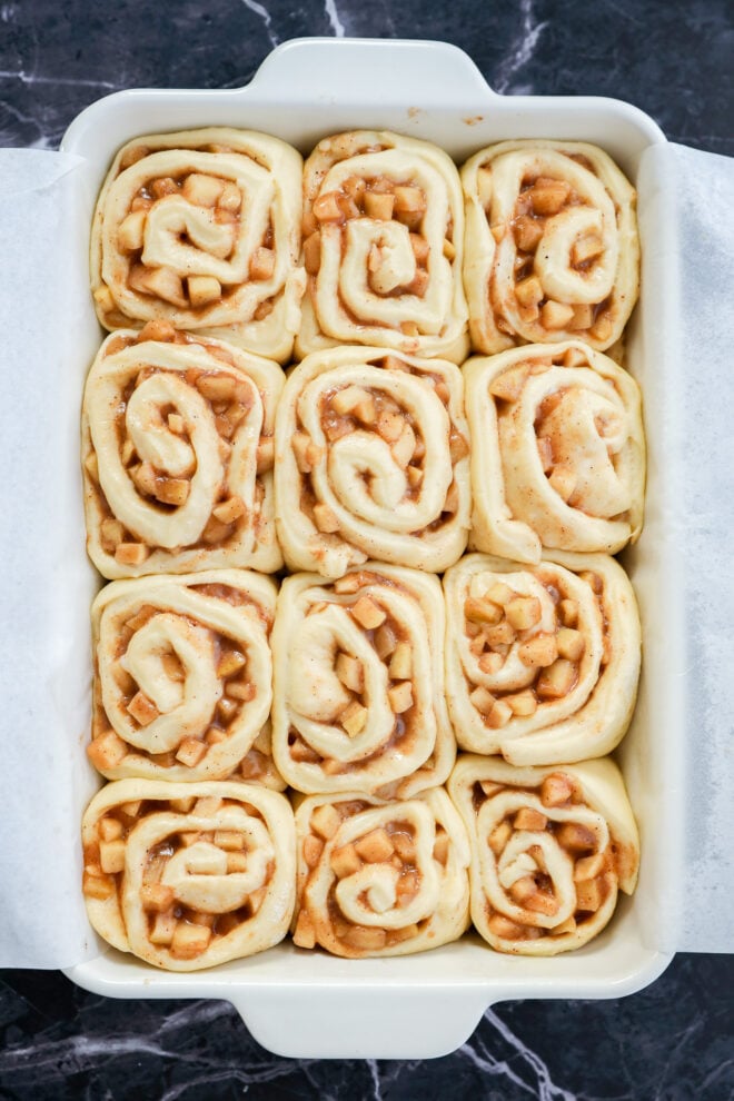 A white baking dish lined with parchment paper holds twelve unbaked Apple Pie Filled Cinnamon Rolls filled with diced apples and cinnamon, arranged in neat rows on a dark marble surface.