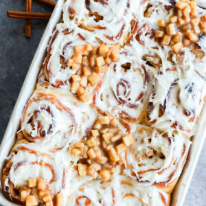 A baking dish filled with Apple Pie Filled Cinnamon Rolls, some topped with small diced apple pieces. Two green apples and cinnamon sticks are in the background on a gray surface.