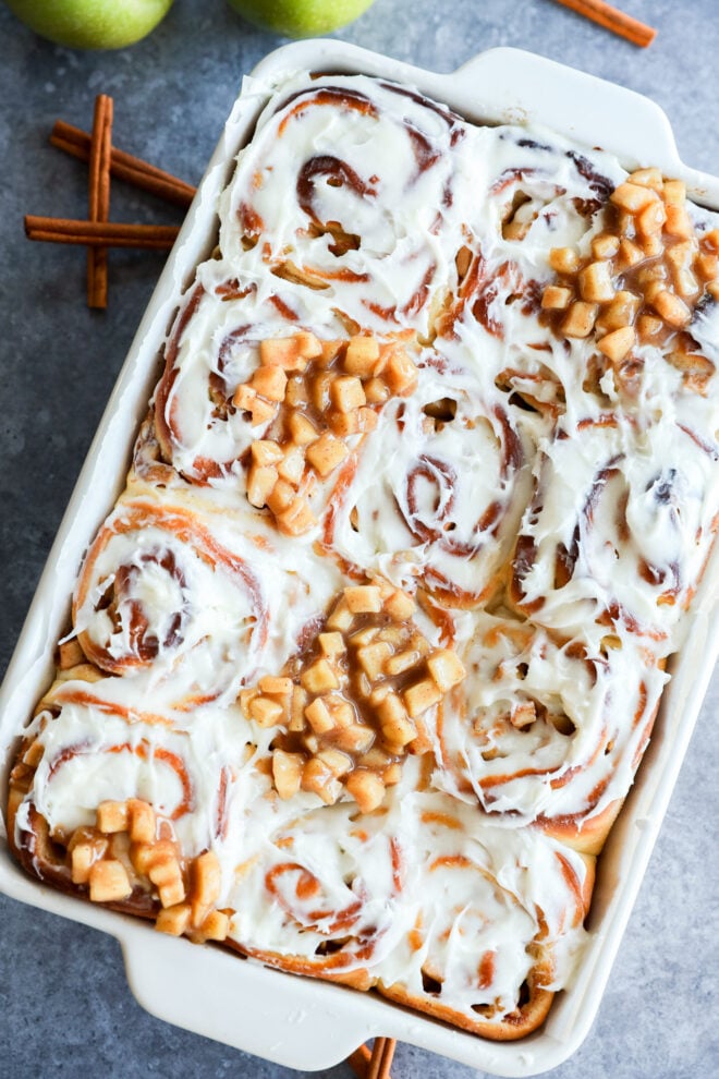 A baking dish filled with Apple Pie Filled Cinnamon Rolls, some topped with small diced apple pieces. Two green apples and cinnamon sticks are in the background on a gray surface.