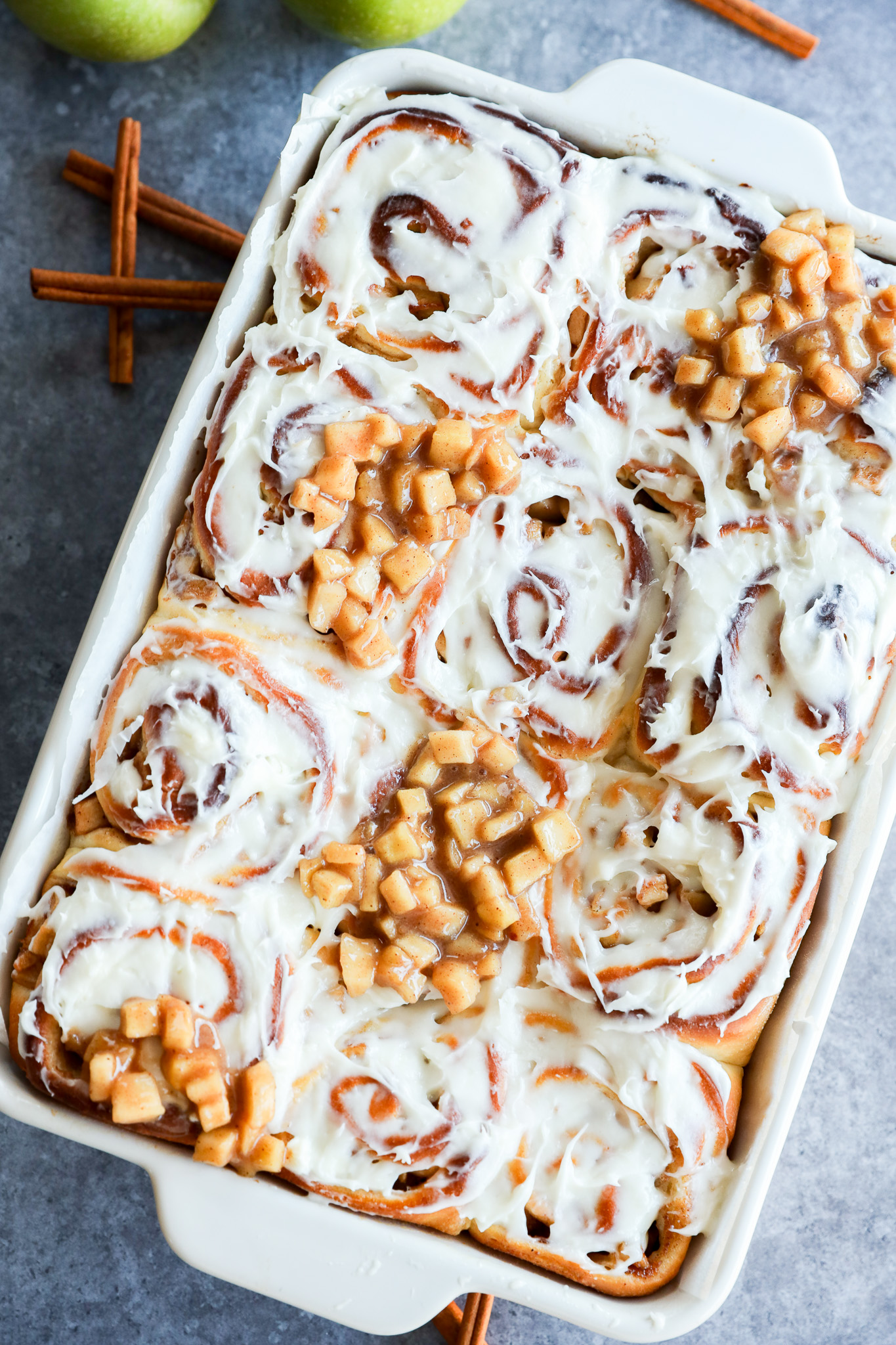 A baking dish filled with Apple Pie Filled Cinnamon Rolls, some topped with small diced apple pieces. Two green apples and cinnamon sticks are in the background on a gray surface.
