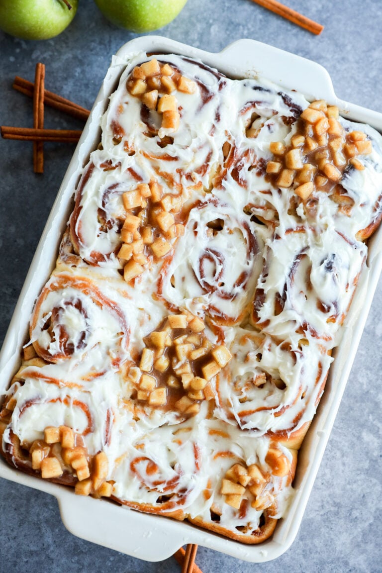 A baking dish filled with Apple Pie Filled Cinnamon Rolls, frosted and topped with small diced apples; cinnamon sticks and green apples are nearby on the gray surface.