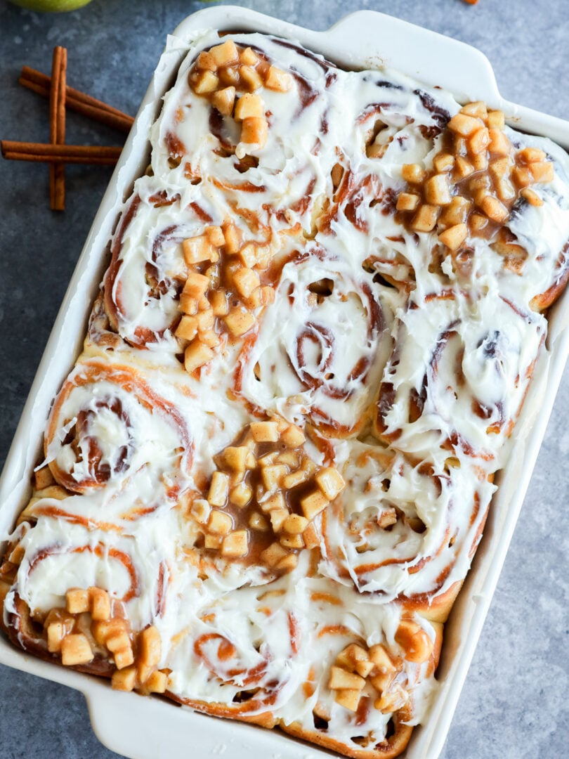 A baking dish filled with Apple Pie Filled Cinnamon Rolls, frosted and topped with small diced apples; cinnamon sticks and green apples are nearby on the gray surface.
