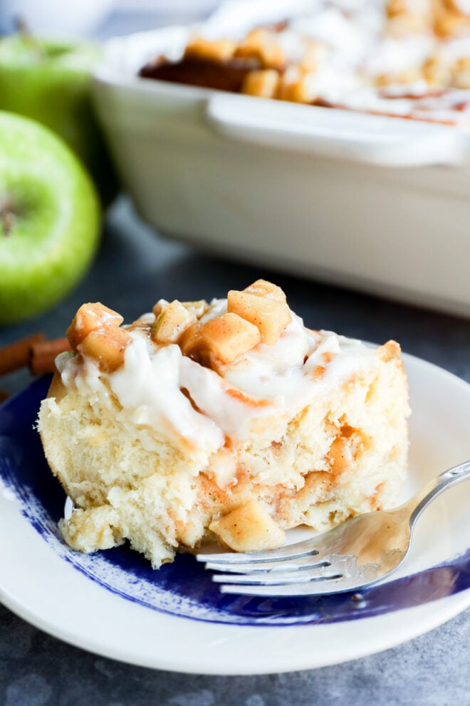 A close-up of Apple Pie Filled Cinnamon Rolls topped with diced apples and white icing on a blue and white plate with a fork. A baking dish and green apples are visible in the background.
