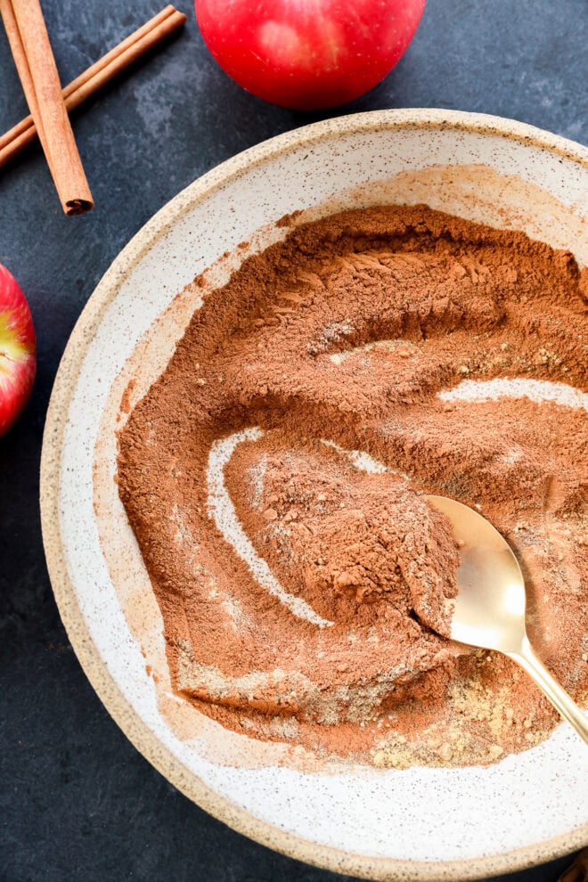 A ceramic bowl filled with a mixture of ground cinnamon and other spices is being stirred with a gold spoon for apple pie spice. Fresh apples and cinnamon sticks are nearby on a dark countertop.