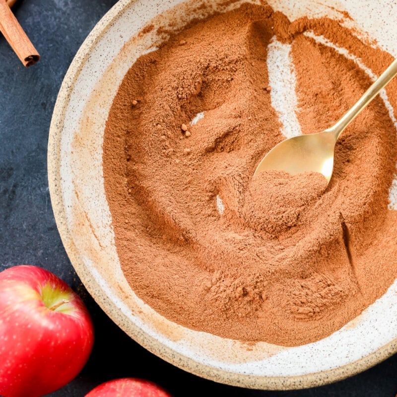 A bowl of apple pie spice with a gold spoon, surrounded by whole red apples and cinnamon sticks on a dark surface.