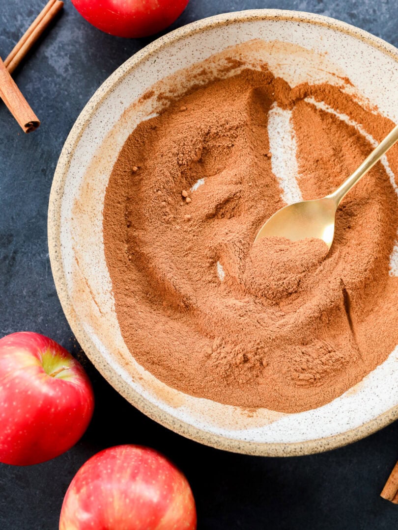 A bowl of apple pie spice with a gold spoon, surrounded by whole red apples and cinnamon sticks on a dark surface.