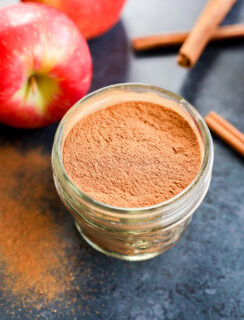 A small glass jar filled with apple pie spice sits on a dark countertop, surrounded by fresh apples and cinnamon sticks.