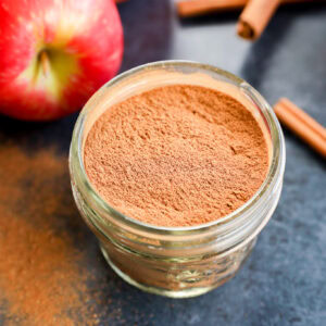 A small glass jar filled with apple pie spice sits on a dark countertop, surrounded by fresh apples and cinnamon sticks.