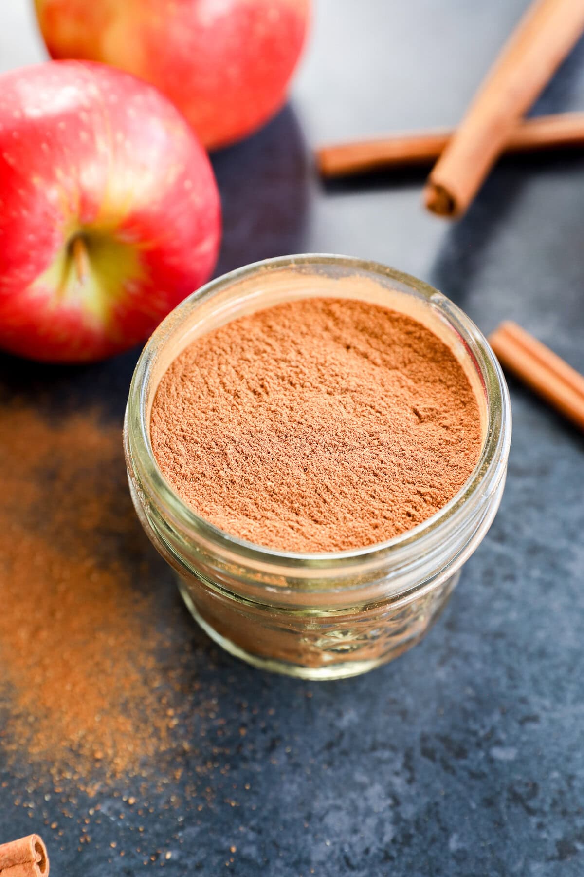 A small glass jar filled with apple pie spice sits on a dark countertop, surrounded by fresh apples and cinnamon sticks.