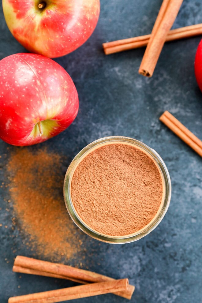 A glass jar filled with a spice mixture sits on a dark surface, surrounded by whole cinnamon sticks, red apples, and scattered cinnamon powder.