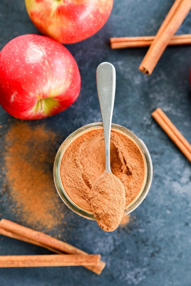 A jar of a spice blend with a spoon rests on top, surrounded by whole red apples and cinnamon sticks on a dark surface. Some cinnamon powder is spilled beside the jar.