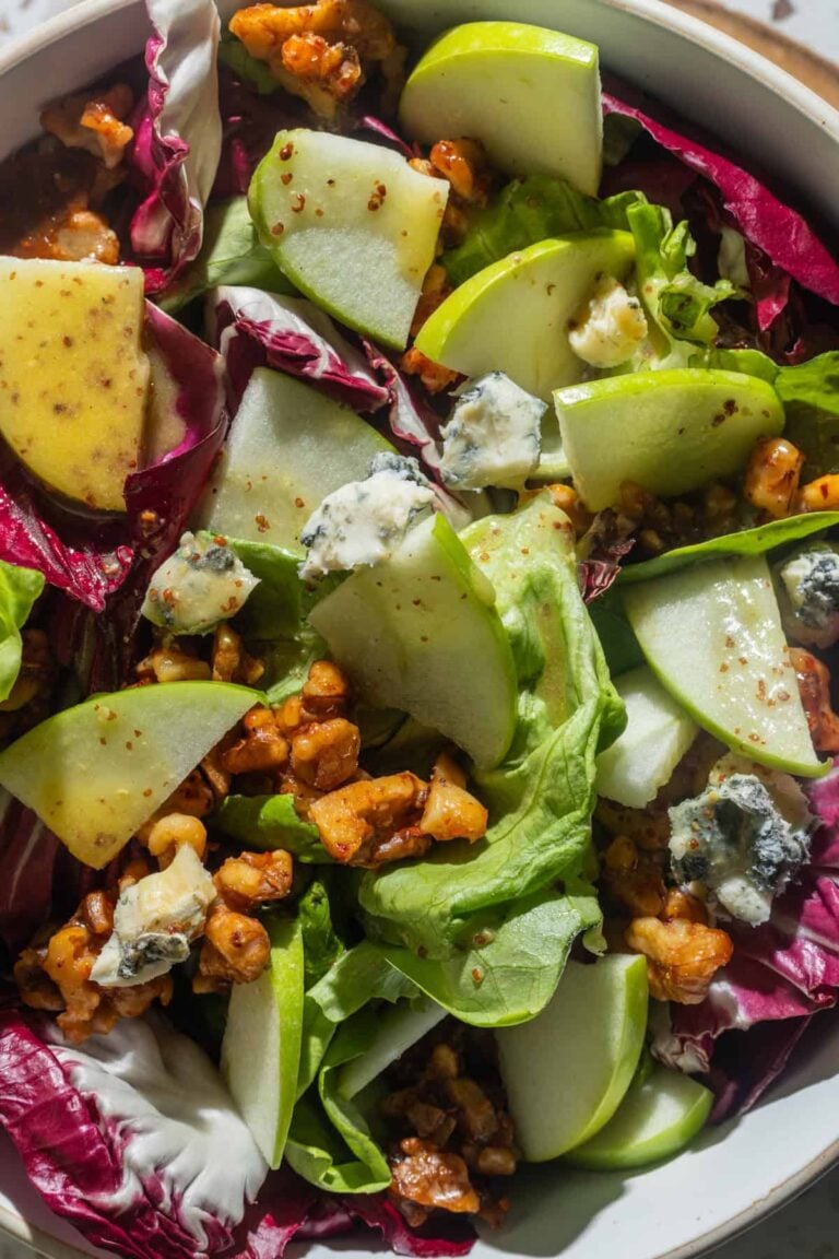 A close-up of a salad with green apple slices, mixed greens, radicchio, crumbled blue cheese, candied walnuts, and a grainy mustard vinaigrette.