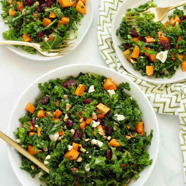 A kale salad with roasted sweet potatoes, dried cranberries, feta cheese, and chopped nuts served in white bowls with gold forks, set on a white surface with a green and white patterned napkin.