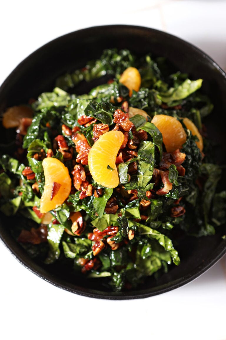 A black bowl filled with a fresh salad of chopped kale, spinach, mandarin orange segments, and candied pecans, set on a white surface. One of the best Thanksgiving salads.