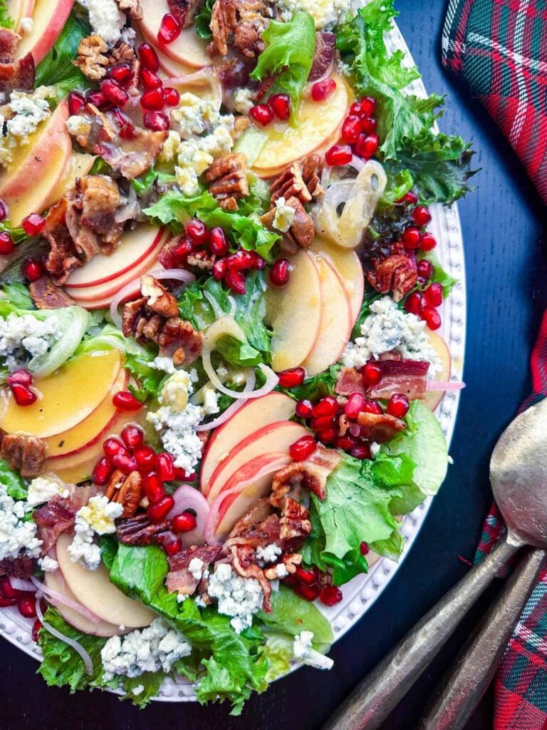 A fresh Thanksgiving salad with sliced apples, crumbled blue cheese, pecans, pomegranate seeds, bacon, and greens on a white plate, next to brass utensils and a red tartan cloth.