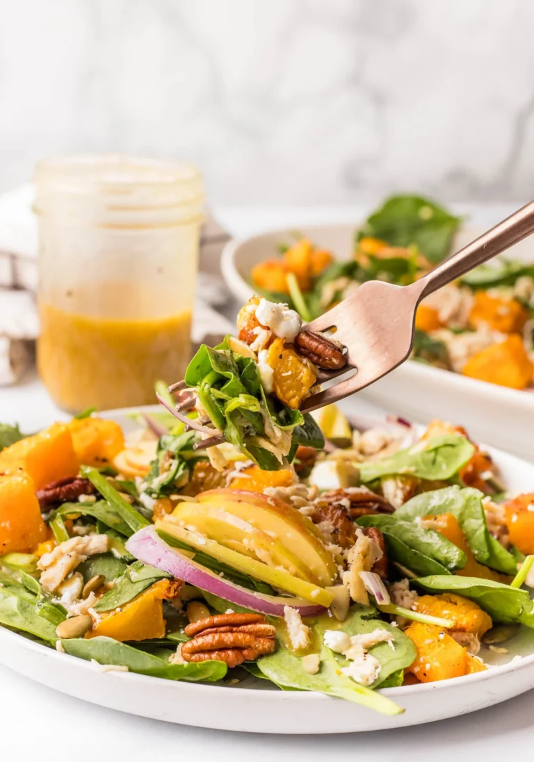 A fork lifts a bite of salad with spinach, roasted butternut squash, red onion, pecans, and sunflower seeds from a plate. A jar of vinaigrette dressing is in the background.