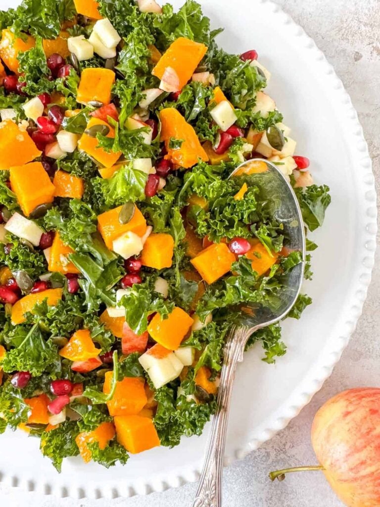 A kale salad on a white plate with chunks of roasted butternut squash, diced apples, pomegranate seeds, and pumpkin seeds. A silver serving spoon rests in the salad. An apple sits nearby on the table.